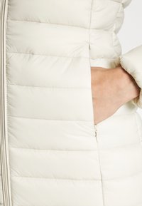 Lys beige polstret jakke med horisontale quiltede linjer, sidelomme med lynlås og elastiske manchetter for en snug pasform.