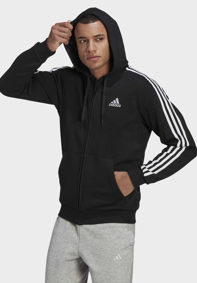 Fato de capuz preto com fecho, com capuz, detalhes em branco nas mangas, bolso canguru, tecido macio, logótipo no peito. Combinado com calças de treino cinzentas.