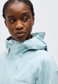 Γυναίκα με πλεγμένα μαλλιά που φοράει ένα ανοιχτό γαλάζιο μπουφάν Columbia με κουκούλα, κοιτάζοντας προς τα αριστερά της, μπροστά σε ένα απλό φόντο.