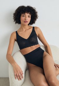 Zwarte kanten bralette met een scalloped rand en dikke banden, gecombineerd met hoge zwarte slips. Gladde textuur, minimalistisch ontwerp.