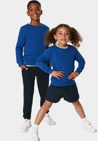 Due bambini stanno insieme, indossando felpe blu coordinate, con pantaloni scuri e scarpe da ginnastica bianche. La ragazza indossa pantaloni corti e calze bianche.