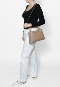 Borsa a mano in pelle marrone con una forma strutturata, manici corti e tracolla. Indossata da una persona con pantaloni bianchi e una maglietta nera.