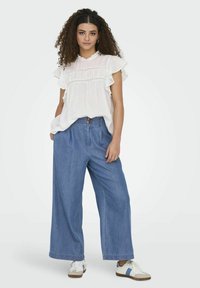 Blusa blanca con mangas acampanadas, lunares texturizados y detalles de encaje, combinada con pantalones de mezclilla de pierna ancha en azul claro y zapatillas blancas.