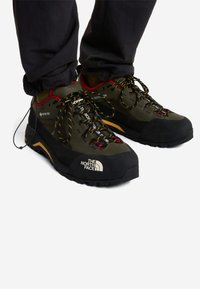 Zapatos de senderismo GORE-TEX en verde oliva y negro, con cordones amarillos y rojos, suela de caucho y detalle del logo de la marca en el lateral.