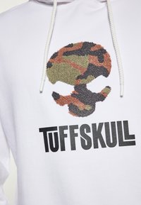 Fehér kapucnis pulóver, zöld, fekete és barna színű, bolyhos katonai álcázású koponya mintával, alatt pedig vastag fekete "TUFFSKUL" felirattal.