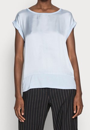 Blouse bleu clair à manches courtes en tissu lisse. Encolure bateau, coupe décontractée, design en couches avec un dos légèrement plus long. Portée avec un pantalon rayé.