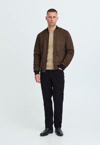 Braune Wildleder-Bomberjacke mit schwarzen gerippten Bündchen und Kragen, kombiniert mit einem beigen Pullover und schwarzen Slim-Fit-Hosen, abgerundet mit schwarzen Stiefeln.