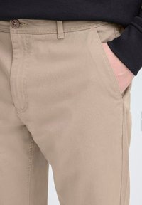 Pantaloni di cotone beige con gamba dritta, dotati di chiusura a bottone, tasca laterale e una texture sottile. Camicia nera a maniche lunghe sullo sfondo.