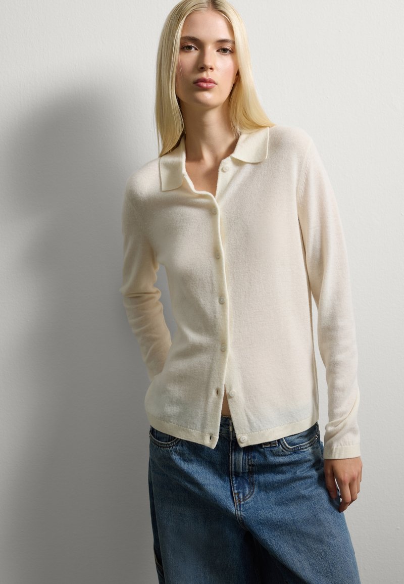 Donna bionda che indossa un cardigan crema con bottoni e jeans blu, in piedi contro un muro grigio chiaro uniforme, con lo sguardo fisso davanti a sé.