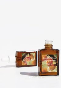 Botella de vidrio marrón de suero de vitamina C con un gotero blanco. La etiqueta presenta gráficos en naranja y verde, y el texto indica 63% de agua de Hippophae Rhamnoides.