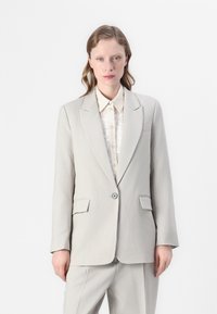 Lys grå skreddersydd blazer med krage, enkel knappelukking og to frontlommer, lagd over en kremfarget skjorte med knapper.