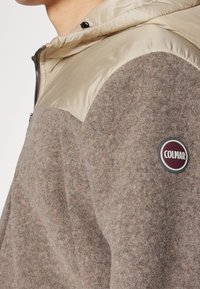 Giacca con cappuccio in pile marrone con pannello sintetico beige sulla spalla, caratterizzata da una toppa con il logo rotondo Colmar sul braccio sinistro. Dettaglio con cerniera visibile.