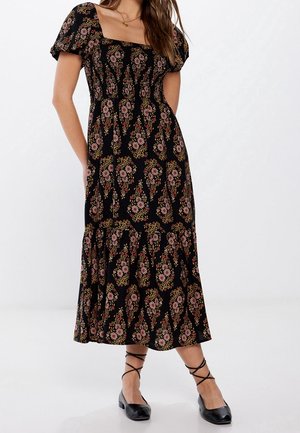 Femme portant une robe midi noire à motif floral rose, manches bouffantes, encolure carrée, et chaussures plates noires à lacets, debout devant un fond blanc.
