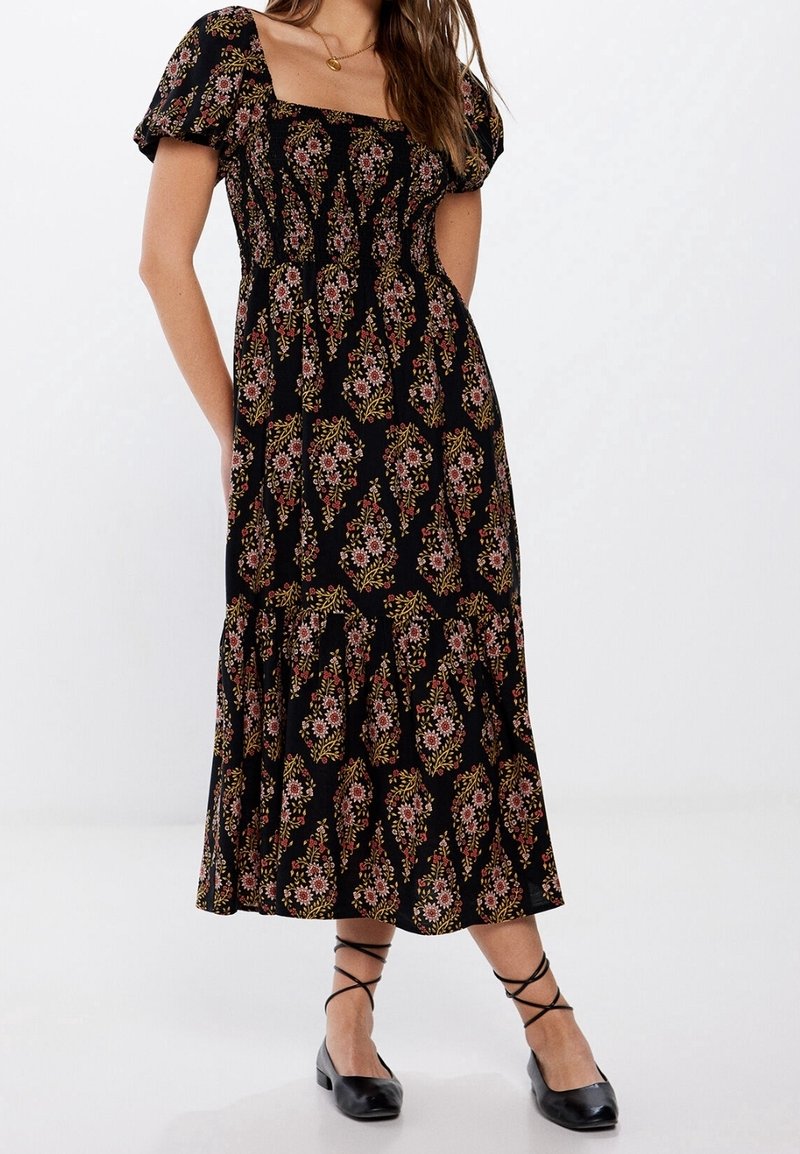 Femme portant une robe midi noire à motif floral rose, manches bouffantes, encolure carrée, et chaussures plates noires à lacets, debout devant un fond blanc.