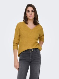 Senfgelber, gerippter Strickpullover mit V-Ausschnitt, lockerer Passform und hochgekrempelten Ärmeln. Kombiniert mit dunkelgrauen Jeans und einem schwarzen Gürtel.