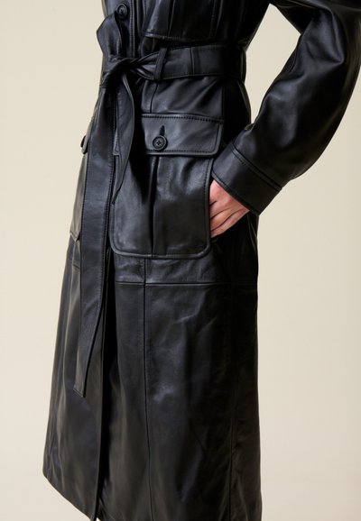 Manteau en cuir noir avec une taille ceinturée, poches boutonnées et coutures détaillées. Texture lisse et manches longes avec une coupe classique.