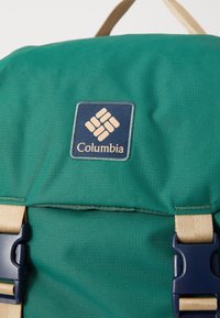 Zaino verde realizzato in tessuto testurizzato, con un patch blu del logo, spallacci beige e clip in blu.