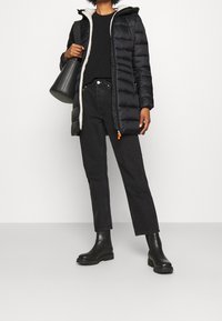Person med sort puffertjakke med hvid kant, sort sweater, højtaljede sorte jeans og sorte ankelstøvler, som holder en sort håndtaske.
