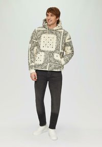 Cremfarbener Kapuzenpullover mit schwarzen Blumen- und geometrischen Mustern, ausgestattet mit einer Fronttasche und gerippten Bündchen, kombiniert mit dunklen Jeans und weißen Turnschuhen.