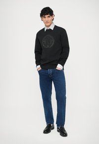 Felpa nera con logo decorato, indossata sopra una camicia azzurra con colletto. Abbinata a jeans blu e scarpe eleganti nere.