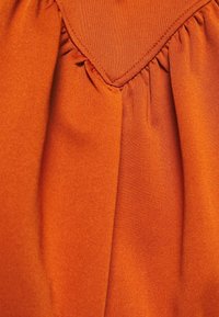 Tela de color óxido con una textura suave, que presenta un diseño fruncido en el escote y un detalle de yugo plano y triangular.