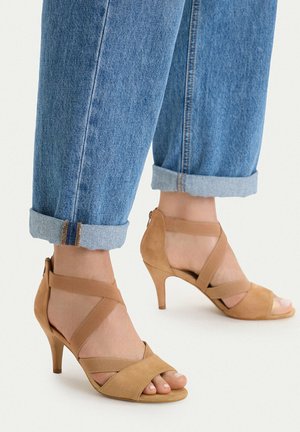 Füße in sandfarbenen Wildleder-High-Heel-Sandalen mit überkreuzten Riemen, kombiniert mit hochgekrempelten blauen Jeans auf weißem Hintergrund.