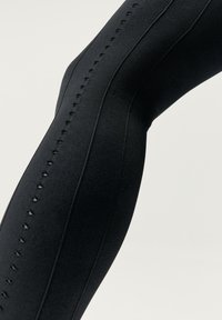 Juodos leggingai su tekstūruotu paviršiumi, vertikaliais siūlais ir dekoratyviais išpjovimais viršutinėje šlaunies dalyje.