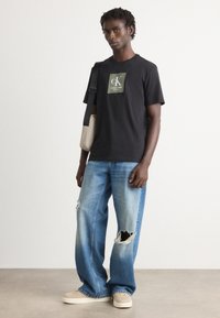 Joven vistiendo una camiseta negra de logo de Calvin Klein, jeans anchos de mezclilla clara con rotos, zapatillas beige y llevando una mochila cruzada.