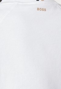 Vit sweatshirt med en mjuk textur, som har en gyllene "BOSS"-logotyp på baksidan, samt en svart och vit randig detalj vid halsringningen.