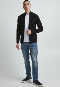 Juodas zip-up džemperis su reljefine tekstūra, dėvimas ant baltos marškinėlių, derinamas su šviesiai mėlynomis džinsomis ir juodais sportbačiais.
