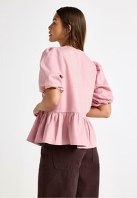 Różowa bluzka z puff-sleeves i peplum, wykonana z teksturowanej tkaniny, w zestawieniu z ciemno-brązowymi spodniami. Prosty design z widokiem z tyłu.