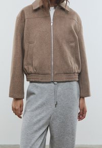 Braune, kurz geschnittene Jacke aus Wollmischung mit Reißverschluss vorne, zwei Seitentaschen und geripptem Saum. Kombiniert mit grauen Jogginghosen mit Kordelzug.