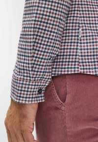 Camicia a quadri in tessuto blu navy, bordeaux e bianco. Presenta polsini con bottoni, bottoni a contrasto e un'etichetta laterale. Indossata con pantaloni in velluto a coste.