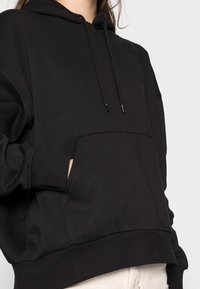 Persona que lleva una sudadera negra con bolsillo frontal y cordones, con una mano parcialmente dentro del bolsillo, combinada con pantalones de color claro.