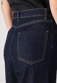 Jeans de denim oscuro con una cintura alta, costuras contrastantes en color marrón claro y dos bolsillos traseros con bordes en ángulo. La textura es suave y resistente.
