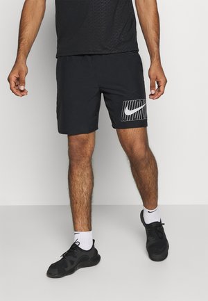 Mężczyzna ubrany w czarne spodenki Nike z białym logo Swoosh, czarne trampki, białe skarpetki Nike oraz czarną teksturowaną koszulkę stoi na szarej podłodze.