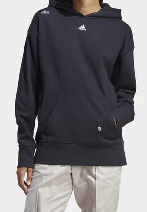 Osoba ubrana w czarną bluzę z kapturem Adidas z przednią kieszenią oraz jasne spodnie, stojąca z jedną ręką w kieszeni, a drugą swobodnie opuszczoną.