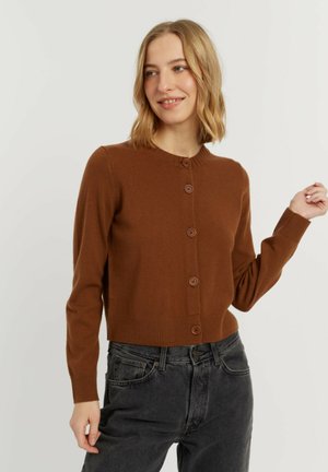 Vrouw met schouderlang blond haar, een bruin cardigan met knopen en een zwarte high-waisted jeans, glimlachend en opzij kijkend.