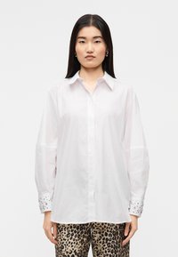 Λευκό πουκάμισο με μακριά μανίκια και κλασικό γιακά, με διακοσμητικά μανσέτα και ίσια ραφή, φορεμένο πάνω από παντελόνι με λεοπάρ σχέδιο.