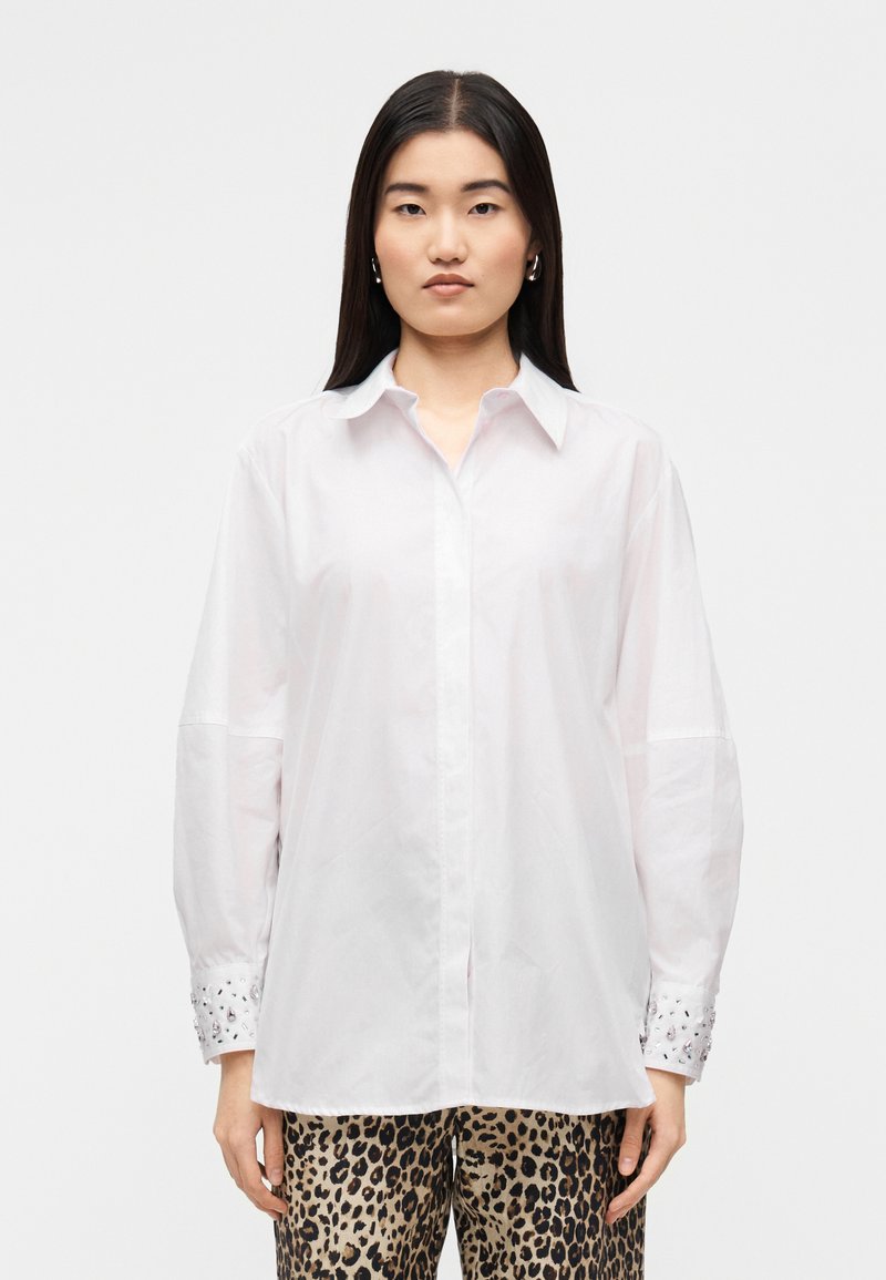 Λευκό πουκάμισο με μακριά μανίκια και κλασικό γιακά, με διακοσμητικά μανσέτα και ίσια ραφή, φορεμένο πάνω από παντελόνι με λεοπάρ σχέδιο.
