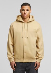 Beige hoodie met een knopenfront, touwtjes en ribbelmanchetten. Draagt over donkere spijkerbroek. Glad materiaal, casual ontwerp.