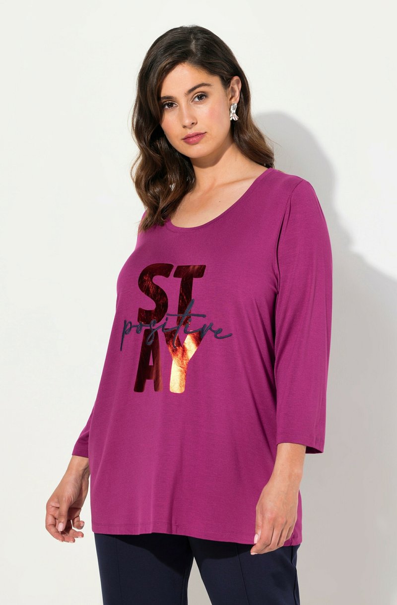Frau mit welligem dunklem Haar, die silberne Ohrringe und ein lila langärmliges Oberteil trägt, auf dem in glänzenden Buchstaben "STAY positive" steht.