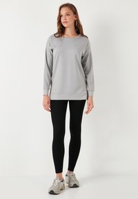 Svijetlosiva dugosleeve pulover s okruglim izrezom, u kombinaciji s crnim tajicama i sivim tenisicama. Glatka tkanina. Minimalistički dizajn.