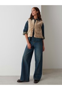 Gilet en fausse fourrure beige porté sur une chemise en denim, associé à un pantalon en jean bleu foncé à jambes larges et des bottes marron. Design uni sans motifs visibles.