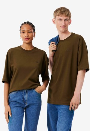 Due giovani adulti che indossano T-shirt marroni oversize e jeans blu, uno con una giacca blu sulla spalla.
