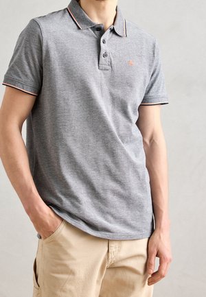 Hombre que lleva una camiseta polo gris de manga corta con ribete naranja y pantalones beige, de pie con una mano en el bolsillo.