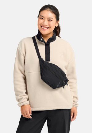 Lächelnde Frau trägt hellbeige Fleece mit schwarzem Kragen, schwarze Umhängetasche mit Reißverschlüssen und schwarze Hose, steht vor weißem Hintergrund.