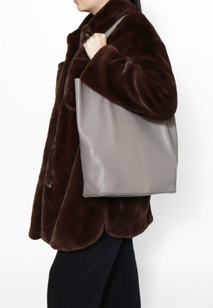 Borsa a tote in pelle grigia con una forma strutturata, spallacci piatti a contrasto e una texture liscia. Indossata sul braccio sopra un cappotto di pelliccia marrone scuro.