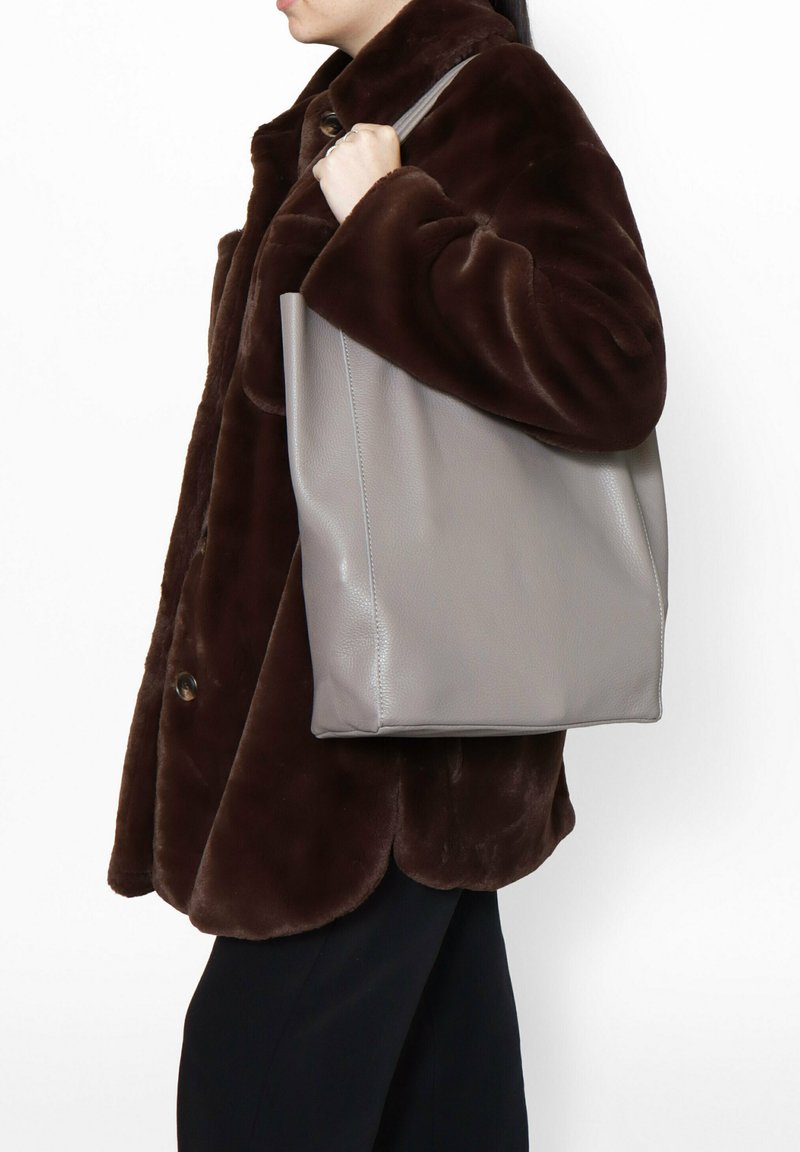 Borsa a tote in pelle grigia con una forma strutturata, spallacci piatti a contrasto e una texture liscia. Indossata sul braccio sopra un cappotto di pelliccia marrone scuro.