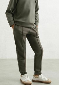 Olivenfarvet, tætsiddende sweatshirt og matchende taperede bukser i blødt stof. Accentueret med sidelommer. Bæres med hvide og beige sneakers.